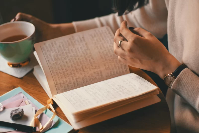 a imagem mostra uma pessoa abrindo um diário com várias escritas sobre a mesa, além de cristais, uma xícara e outro itens