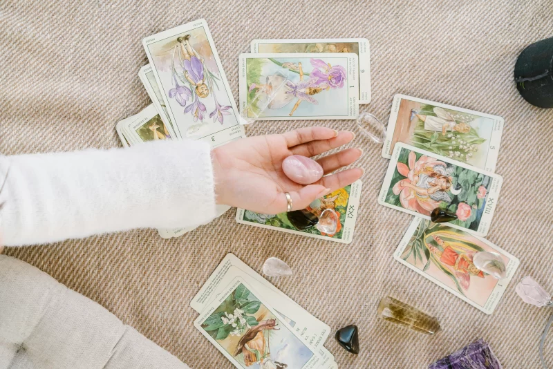 a imagem mostra uma mão segurando um cristal quartzo rosa com cartas de tarot sobre o tapete para entender como aprender a ler tarot e fazer uma tiragem pessoal