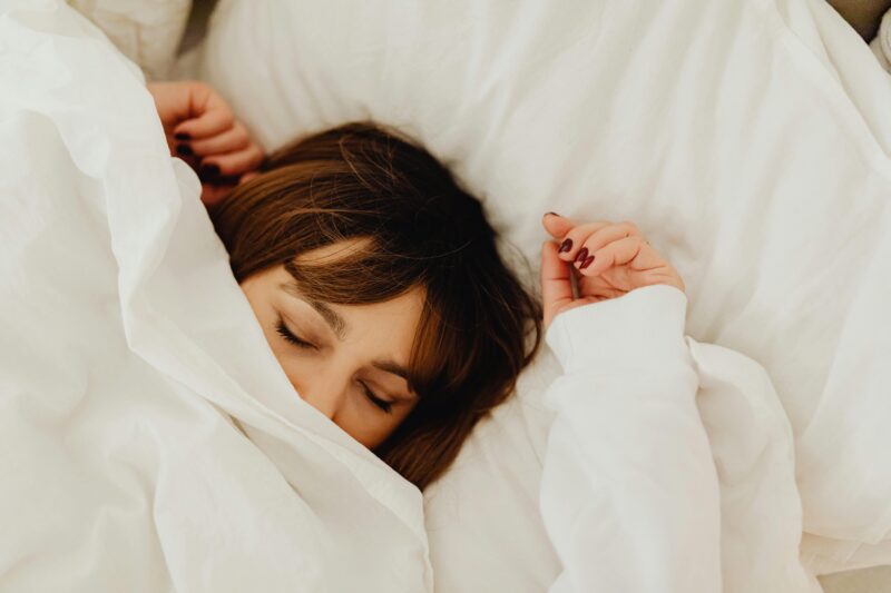 a imagem mostra uma mulher coberta com o lençol aparecendo só os olhos, cabelo, parte do nariz e as mãos, descansando na cama