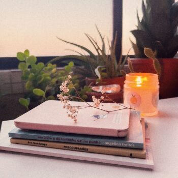 livros de poesia sobre a mesa, com ramo de flores, vela aromática acesa e plantas abaixo de uma janela de apartamento
