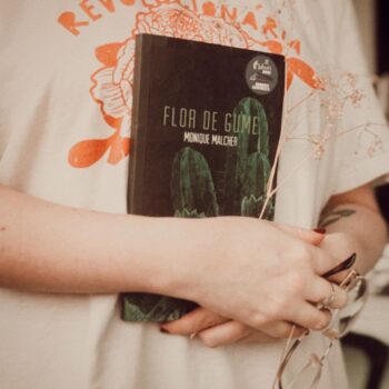 o livro mostra uma pessoa segurando o livro Flor de Gume, da escritora Monique Malcher, mostrando a capa da obra, um óculos de grau e um ramo de flores secas